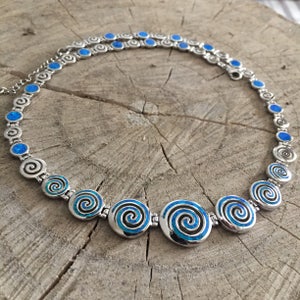 May include: A silver necklace with blue opal swirl pendants. The necklace is made of a chain of small silver circles with blue opal swirls inside. The necklace has a larger pendant in the center with a blue opal swirl.