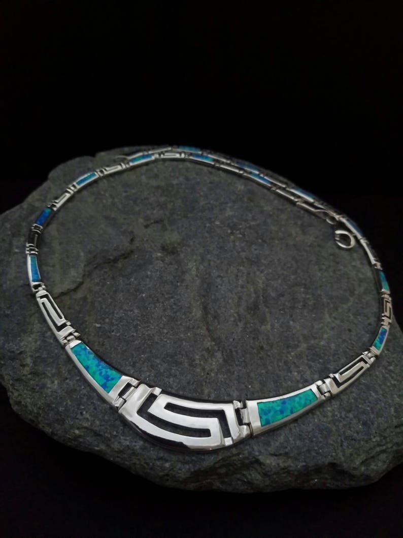 May include: A silver necklace with a geometric design and blue opal accents. The necklace is on a gray rock.