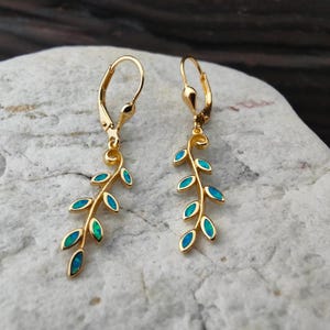 May include: A pair of gold-colored earrings featuring a leaf design. Each earring has a lever back closure and a stem with several blue-green oval-shaped accents, resembling leaves. The earrings are displayed on a light-colored, textured surface.