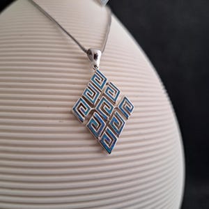 May include: A silver necklace with a blue and silver pendant featuring a geometric design. The pendant is shaped like a diamond and has a repeating pattern of interlocking squares.