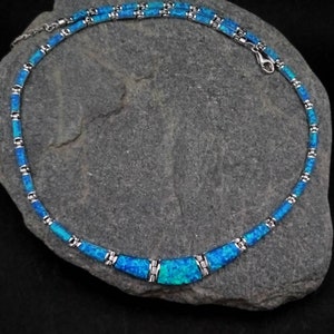Puede incluir: Un collar de plata con una pieza central de &oacute;palo azul. El collar est&aacute; hecho de peque&ntilde;os cuadrados de plata unidos con incrustaciones de &oacute;palo azul. El collar est&aacute; sobre un fondo de piedra gris.