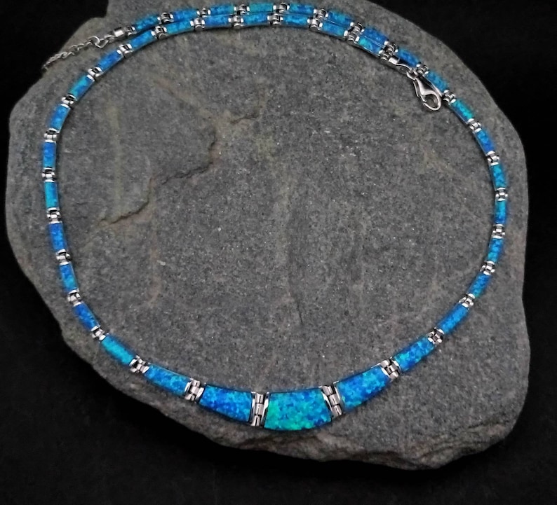 Puede incluir: Un collar de plata con una pieza central de &oacute;palo azul. El collar est&aacute; hecho de peque&ntilde;os cuadrados de plata unidos con incrustaciones de &oacute;palo azul. El collar est&aacute; sobre un fondo de piedra gris.
