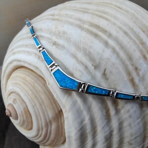 May include: A silver necklace with geometric shapes filled with bright blue opal. The necklace is displayed on a large, textured seashell. The opal has a speckled appearance, and the silver setting has a polished finish. Jewelry.