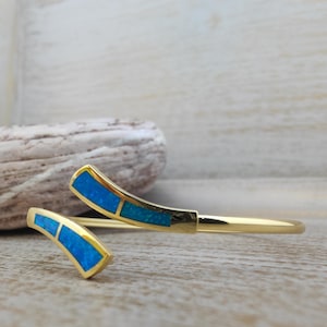 Puede incluir: Una pulsera de brazalete abierto en tono dorado con dos extremos curvos, cada uno incrustado con una piedra azul vibrante estilo ópalo. La banda de metal pulido tiene un acabado liso y reflectante, creando un espacio entre los dos segmentos.