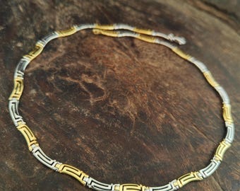 May include: A gold and silver-toned necklace with a geometric design. The necklace is composed of alternating gold and silver segments, creating a striking contrast. The necklace is displayed on a dark wooden surface.