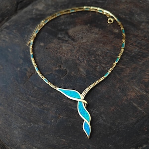 May include: Gold-toned necklace with a turquoise blue pendant. The necklace features a delicate chain with turquoise accents and a stylized, intertwined pendant. The design is set against a dark, textured background, highlighting the jewelry's intricate details.