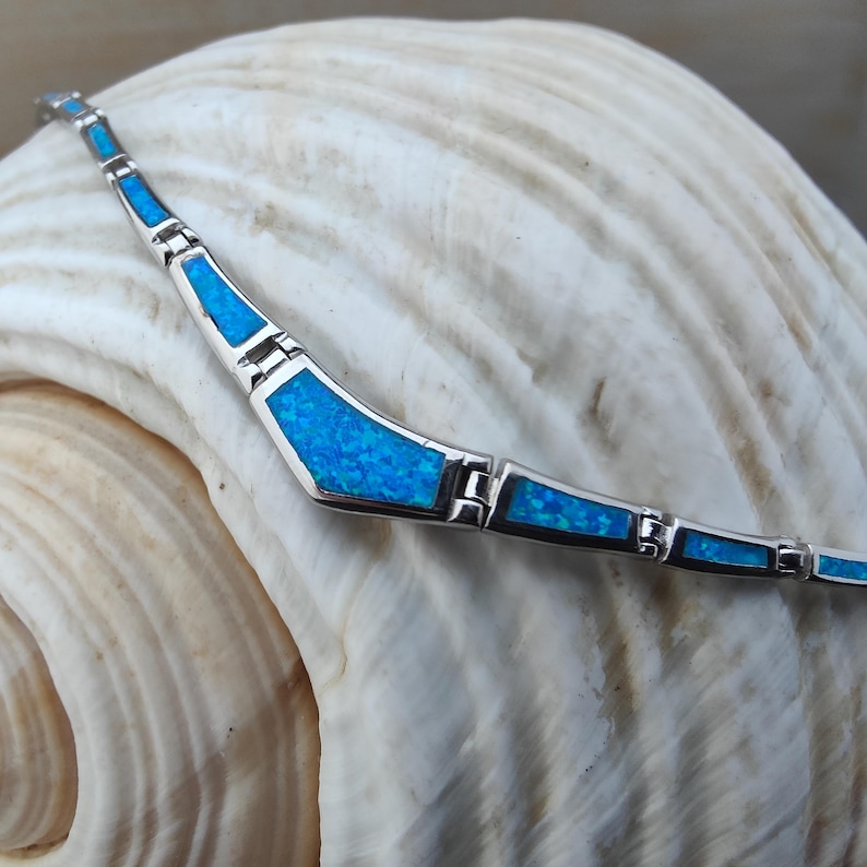 May include: A silver bracelet with blue opal inlays. The bracelet features a series of geometric shapes filled with vibrant blue opal, connected by silver links. The bracelet is displayed on a white seashell.