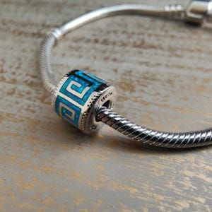 Könnte beinhalten: Ein silberner Charm-Bead mit einem geometrischen Design in Blau- und Weißtönen. Der Bead ist zylindrisch und weist ein sich wiederholendes Muster auf. Der Charm befindet sich an einem silbernen Schlangenkettenarmband.