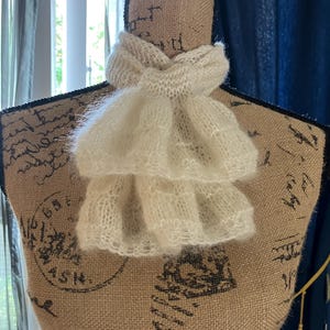 May include: A cream-colored, knitted neck ruff, displayed on a mannequin. The ruff features a layered, ruffled design, adding a touch of vintage elegance. The soft, textured knit creates a delicate and airy appearance. The mannequin is covered in a burlap-like fabric.