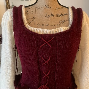 Puede incluir: Un corpiño de punto burdeos con escote cuadrado y frente con cordones, combinado con una camiseta blanca de manga larga. El corpiño tiene un dobladillo con volantes y se exhibe en un maniquí.