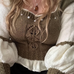 May include: A brown and white knitted sweater with a pearl trim and a ruffled hem. The sweater has a scoop neck and long sleeves.
