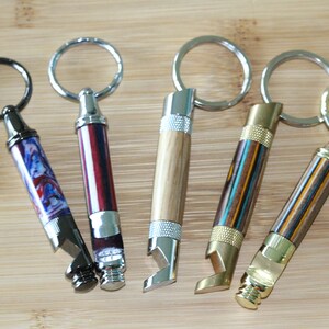May include: Six different bottle opener keychains with various wood and metal finishes. Each keychain has a different wood pattern and metal accents. The keychains are all in focus and arranged in a row on a wooden surface.