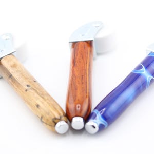 May include: Three wooden and resin roller handles with white wheels. The handles are made of different types of wood and resin, including a brown wood with dark streaks, a reddish-brown wood, and a blue resin with white swirls.