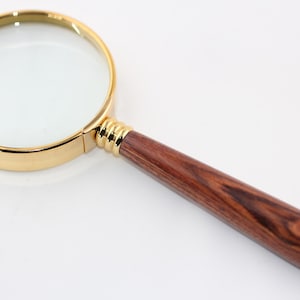 May include: A magnifying glass featuring a circular lens encased in a polished gold-tone metal frame. The handle is crafted from dark brown wood with a distinct natural grain pattern, accented by gold-tone metal rings at the base of the lens.