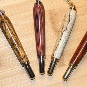 May include: Four wooden pen-shaped pendants with gold-toned caps and chains. The pendants are made of different types of wood with unique grain patterns.