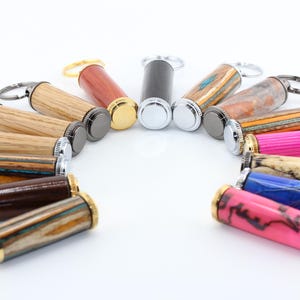 May include: A collection of cylindrical keychains arranged in a semi-circle. The keychains feature various materials including natural wood grains, polished resins, and vibrant pink, blue, and multi-colored patterns with gold, silver, and black metal hardware.