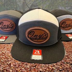 May include: Three black, gray, and white flat bill snapbacks with a brown leather patch that says "Onyx Solutions" in a cursive font. The hats are on a bed of brown leaves.
