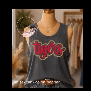 May include: A gray unisex tank top with the word "Tigers" in red script outlined in gold glitter. The tank top is displayed on a mannequin. The text "unisex tank color: pepper" is at the bottom of the image.