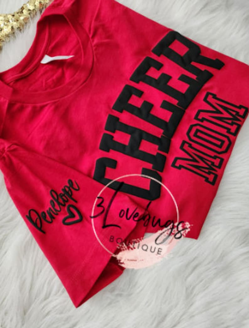 May include: Red t-shirt with black text that says "Cheer Mom" and a smaller black text that says "Penelope" with a heart on the sleeve.
