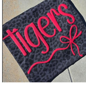 May include: A dark gray decorative pillow with a leopard print pattern. The word "tigers" is spelled out in bright red, cursive letters across the pillow. A red bow is also featured on the pillow.