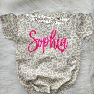 May include: A beige and brown leopard print t-shirt with the name "Sophia" in neon pink puff lettering. The shirt has short sleeves and a crew neck.