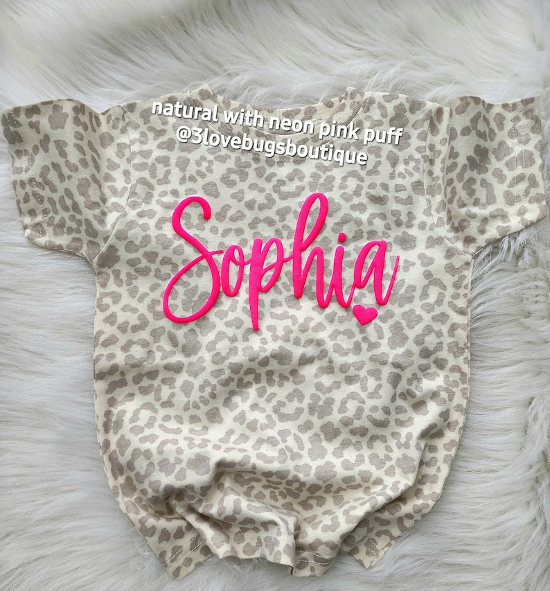 May include: A beige and brown leopard print t-shirt with the name "Sophia" in neon pink puff lettering. The shirt has short sleeves and a crew neck.