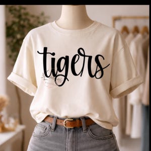Könnte beinhalten: Cremefarbenes T-Shirt mit dem Wort "tigers" in schwarzer Schrift. Das Shirt hat hochgekrempelte Ärmel und wird mit grauen Jeans und einem braunen Gürtel kombiniert. Das T-Shirt wird auf einer Schaufensterpuppe präsentiert.