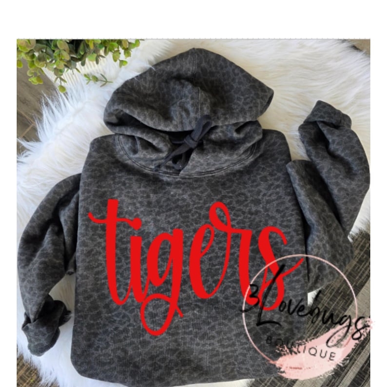 May include: A gray hoodie with a leopard print pattern. The word "tigers" is written in red script across the front. The hoodie has a drawstring hood and long sleeves. The image also includes a white fur-like surface.