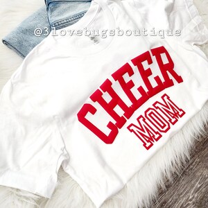 May include: White t-shirt with the words "Cheer Mom" in red block letters.