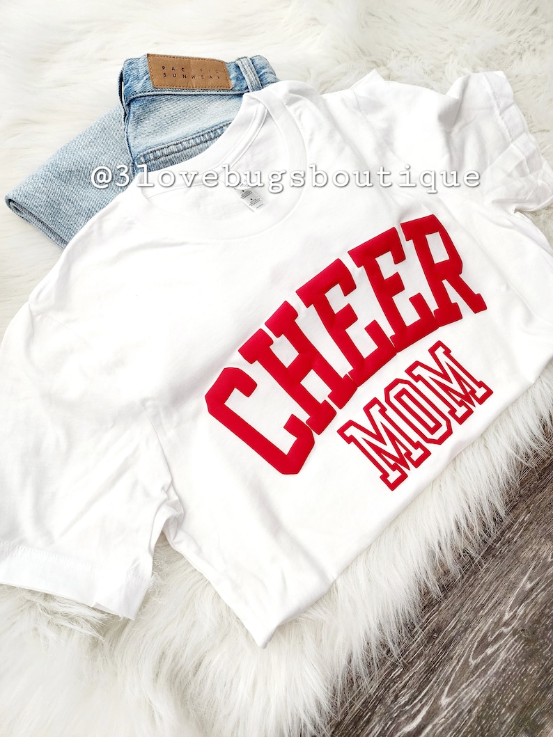 May include: White t-shirt with the words "Cheer Mom" in red block letters.
