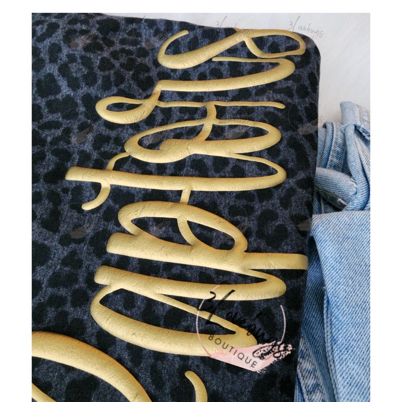 May include: A folded piece of fabric with a dark gray leopard print pattern. The word "Sassy" is written in gold cursive lettering across the fabric. A light blue denim garment is visible in the background.