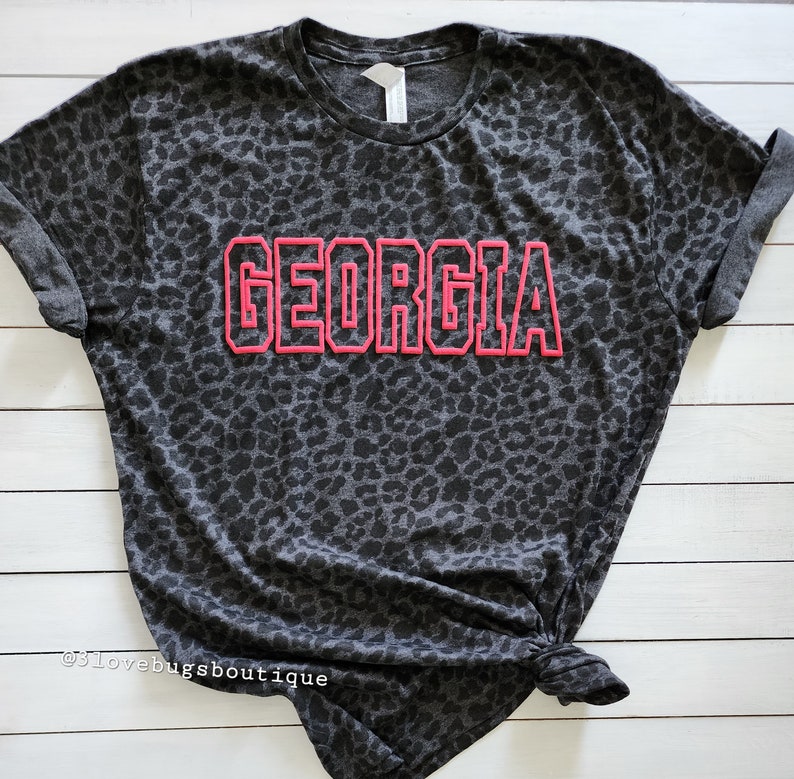 May include: A grey t-shirt with a black leopard print and the word "GEORGIA" in pink outlined letters.