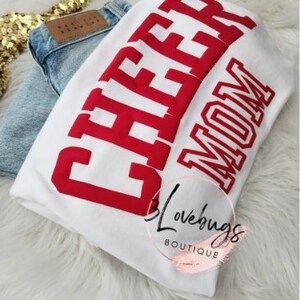 May include: White t-shirt with red text that says "Cheer Mom".