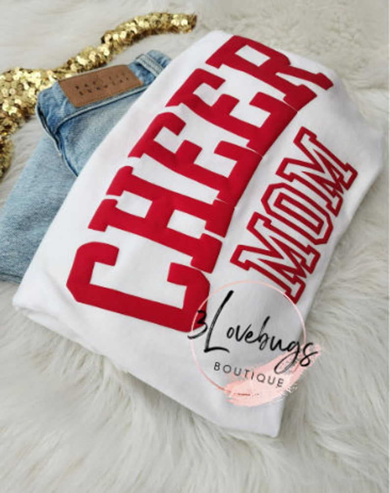 May include: White t-shirt with red text that says "Cheer Mom".