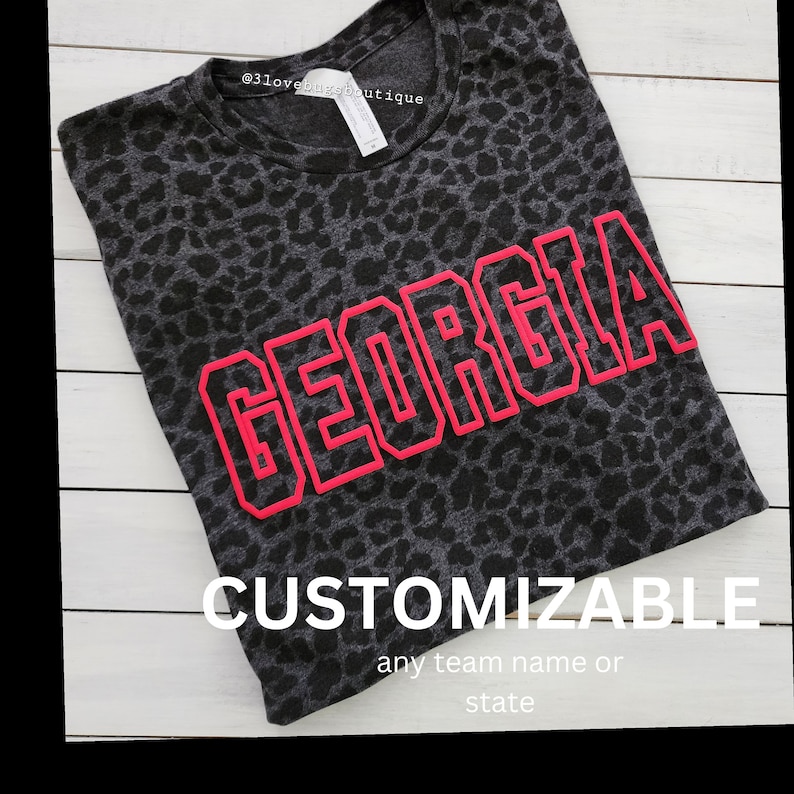 May include: Black and grey leopard print t-shirt with the word "GEORGIA" in large pink letters with a black outline. The text "CUSTOMIZABLE" is below the word "GEORGIA" with the text "any team name or state" below that.