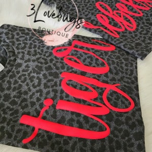 May include: Two gray leopard print t-shirts with the word "Vibe" and "Rebel" in bright red lettering. The shirts are displayed on a white, fluffy surface. The brand name "3 Lovebugs Boutique" is visible in the background.