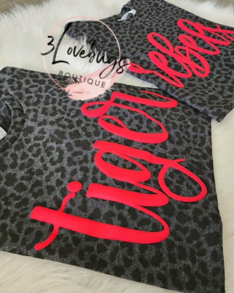 May include: Two gray leopard print t-shirts with the word "Vibe" and "Rebel" in bright red lettering. The shirts are displayed on a white, fluffy surface. The brand name "3 Lovebugs Boutique" is visible in the background.