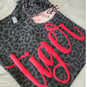 May include: A gray leopard print t-shirt with the word "Tiger" in bright pink, stylized lettering. The shirt has a crew neck and short sleeves. The image also includes the text "B Lovebugs Boutique" in a circular design.