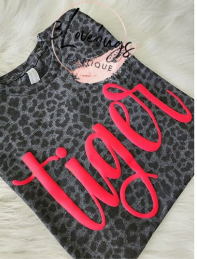 May include: A gray leopard print t-shirt with the word "Tiger" in bright pink, stylized lettering. The shirt has a crew neck and short sleeves. The image also includes the text "B Lovebugs Boutique" in a circular design.