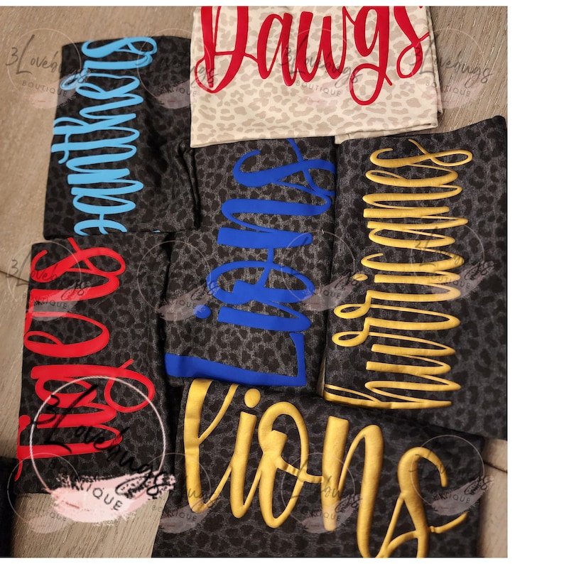 May include: A collection of athletic leggings with various team names in different colors. The leggings have a dark gray leopard print background. The team names include "Panthers", "Dawgs", "Lions", "Hurricanes", and "Saints".