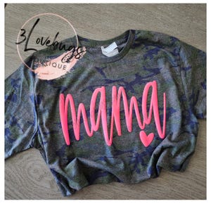 May include: A camouflage-patterned t-shirt with the word "mama" in bright pink script across the front. A small pink heart is next to the word. The shirt has a crew neck and short sleeves.