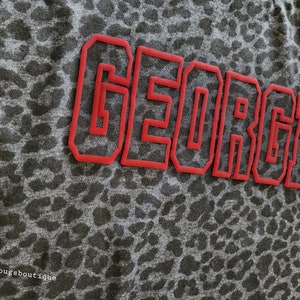 May include: A grey shirt with a leopard print pattern. The word "GEORGIA" is written in red outlined letters across the front of the shirt.