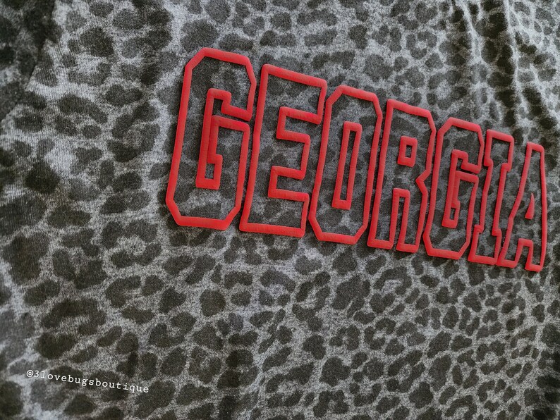 May include: A grey shirt with a leopard print pattern. The word "GEORGIA" is written in red outlined letters across the front of the shirt.