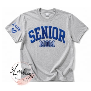 May include: A heather gray short-sleeve t-shirt featuring the blue text SENIOR MOM in a collegiate font on the chest. The right sleeve displays the blue text Laila with a heart symbol. A circular logo with the text 3lovebugs BOUTIQUE is visible.