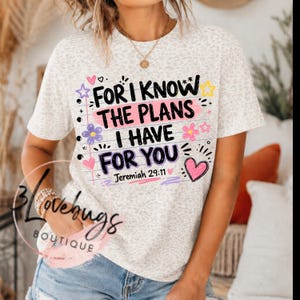 May include: A short-sleeve t-shirt with a light gray leopard print pattern. The front features the black text "FOR I KNOW THE PLANS I HAVE FOR YOU" and "Jeremiah 29:11" surrounded by colorful hand-drawn style hearts, stars, and flowers.