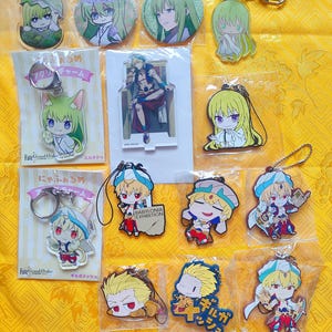 Peut inclure: Une collection de produits dérivés de personnages d'anime, comprenant des porte-clés, des badges et un présentoir. Les articles présentent divers personnages avec des coiffures et des tenues distinctives. Le fond est un tissu jaune vif.