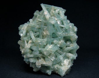 May include: A cluster of pale blue-green aquamarine crystals. The crystals are elongated and form a complex, interwoven structure. The mineral specimen has a rough, textured surface and is set against a dark background.