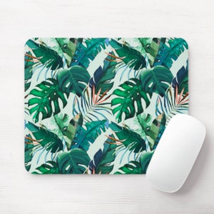 May include: A rectangular mouse mat with a lush tropical leaf design in greens and blues. The pattern showcases large monstera leaves, palm fronds, and other tropical foliage. A white computer mouse is positioned on the right side of the mouse mat.