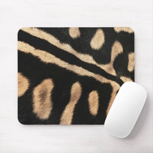 Pode incluir: Um mousepad retangular com um design de estampa de animal preto e castanho. Um mouse de computador branco está posicionado no lado direito do mousepad. O mousepad tem cantos arredondados e está em uma superfície branca.