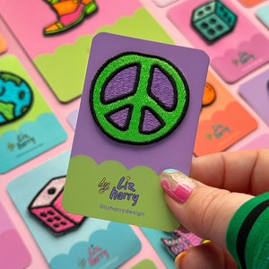 May include: A collection of colourful embroidered iron-on patches featuring a peace sign, a dice, a mushroom, a planet, and a rocket ship. The patches are on a pink background with the text "by Liz harry @lizharrydesign".